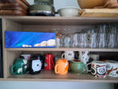 Shelf with various cups, mugs, and glassware in a kitchen setting with a small canvas painting of a mother and cub polar bear.